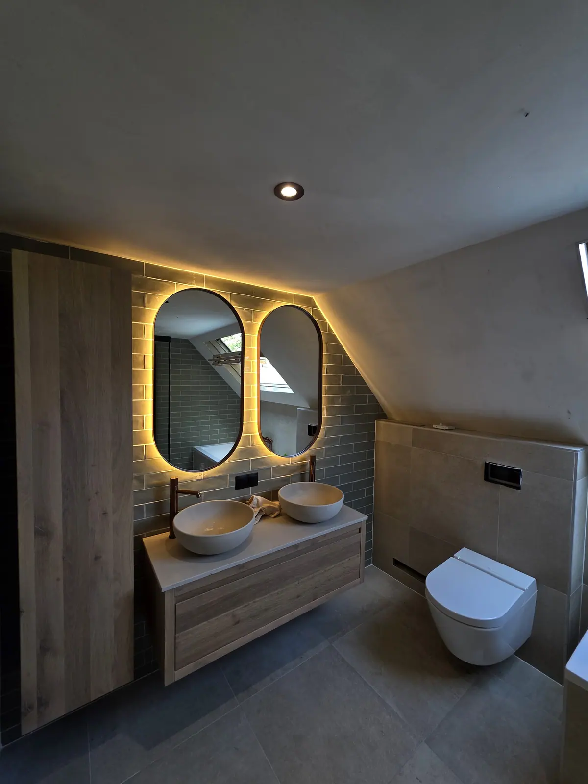 Verbouwde badkamer onder een schuin dak met houten badmeubel, twee ronde ledspiegels en hangtoilet