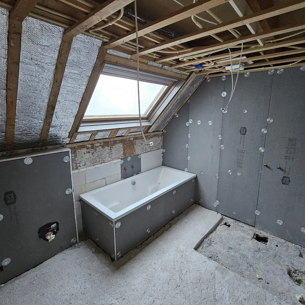 Badkamer onder een schuin dak tijdens de verbouwing, met bad, wandplaten en leidingwerk